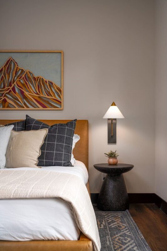 Guest bedroom in the Suncadia home with a camel leather upholstered bed, layered neutral and navy check pillows, a framed mountain landscape painting, brass wall sconce, and a dark pedestal side table.